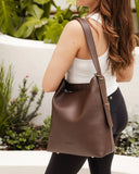 Woman carrying a brown faux leather bag outdoors with greenery in the background