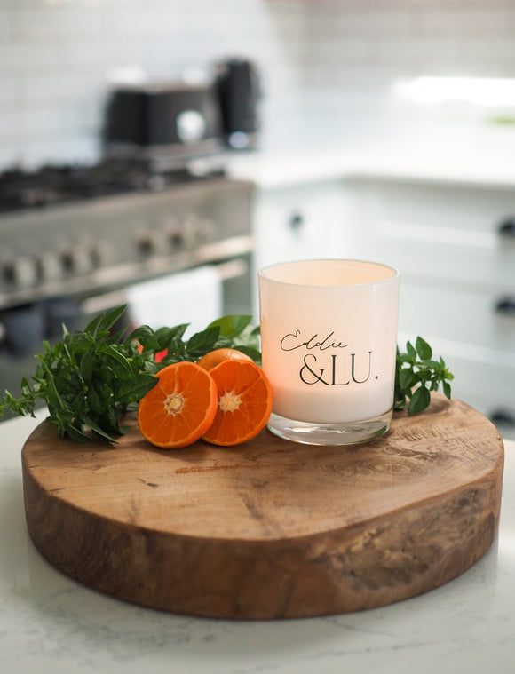 Candle with 'Eldin & Lu' branding on a wooden board with oranges and greenery in a kitchen setting